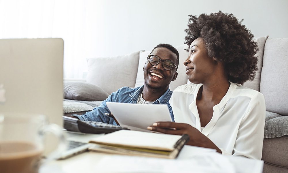Blog - Couple Smiling as they Sit on the Floor in Front of their Couch in their Home as They Review Paperwork and Work on a Laptop