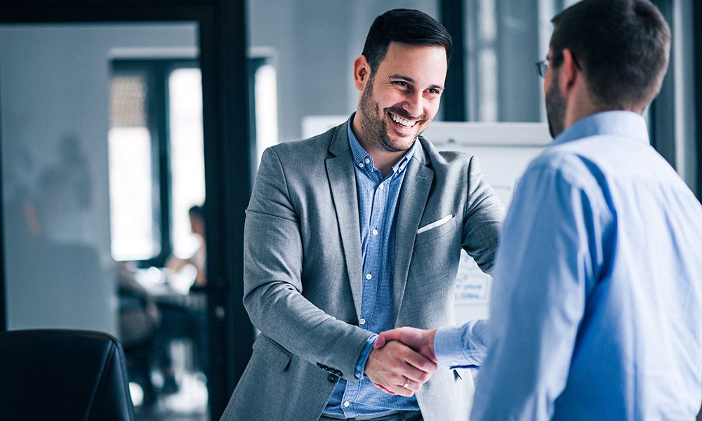 Blog - Two Professional Men Shaking Hands and Smiling in an Office in Front of a Window with Sun Pouring In